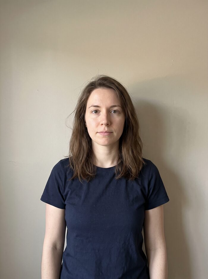 A young woman against a beige wall at home, snapped on a phone from waist up.